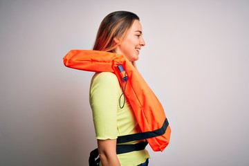 Young beautiful blonde woman with blue eyes wearing orange lifejacket over white background looking...