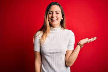 Beautiful blonde woman with blue eyes wearing casual white t-shirt over red background smiling cheerful presenting and pointing with palm of hand looking at the camera.
