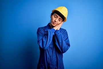 Young beautiful worker woman with blue eyes wearing security helmet and uniform sleeping tired dreaming and posing with hands together while smiling with closed eyes.