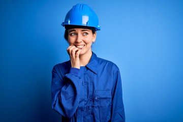 Young beautiful worker woman with blue eyes wearing security helmet and uniform looking stressed and nervous with hands on mouth biting nails. Anxiety problem.