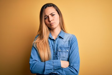 Young beautiful blonde woman with blue eyes wearing denim shirt over yellow background skeptic and nervous, disapproving expression on face with crossed arms. Negative person.