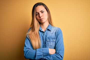 Fototapeta premium Young beautiful blonde woman with blue eyes wearing denim shirt over yellow background skeptic and nervous, disapproving expression on face with crossed arms. Negative person.