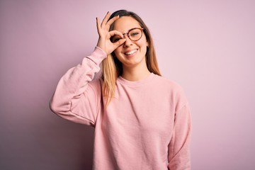 Beautiful blonde woman with blue eyes wearing sweater and glasses over pink background doing ok gesture with hand smiling, eye looking through fingers with happy face.