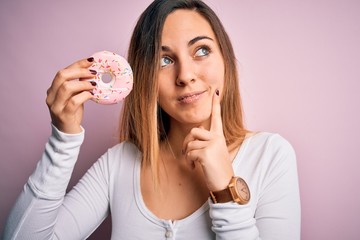 Young beautiful blonde woman with blue eyes holding pink doughnut over isolated background serious...