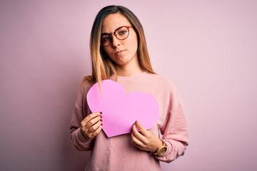 Young beautiful blonde romantic woman wearing glasses holding big pink heart with a confident expression on smart face thinking serious