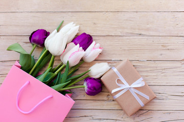 Spring holidays. Tulips and gift box at wooden background. Blooming flowers in pink shopping bag. White, lilac and purple bouquet. Flat lay. Copy space.