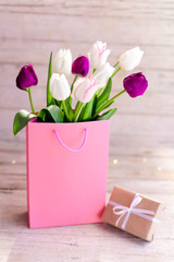 Spring gift. Tulips and box with present at wooden background. Blooming flowers in pink shopping bag. White, lilac and purple bouquet. Still life in morning sun light.