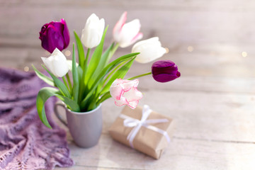 Spring flowers and gift box at wooden background. Blooming tulips in gray cup. Pink, white, lilac and purple bouquet in vase. Cozy still life. Copy space.