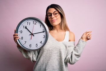Young beautiful brunette woman wearing glasses doing countdown using big clock very happy pointing...