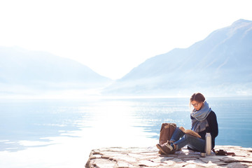 Young woman reading book at sea beach. Cozy winter picnic by morning mountains. Girl in blue scarf enjoying traveling. Traveler drinks coffee. Solo female tourism. Lifestyle moment. Copy space.