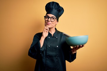 Young beautiful brunette chef woman wearing cooker uniform and hat holding bowl and whisk serious...