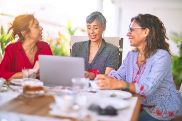 Meeting of middle age women having lunch and drinking coffee. Mature friends smiling happy using laptop at home on a sunny day