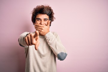 Young handsome man wearing casual t-shirt standing over isolated pink background laughing at you, pointing finger to the camera with hand over mouth, shame expression