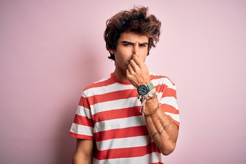 Young handsome man wearing striped casual t-shirt standing over isolated pink background smelling...