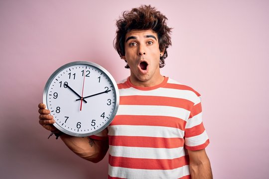 Young handsome man holding big clock standing over isolated pink background scared in shock with a surprise face, afraid and excited with fear expression