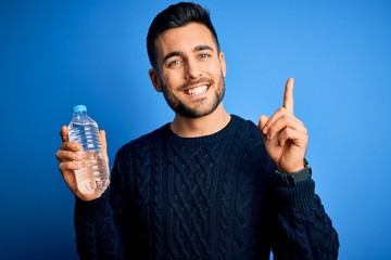 Young handsome man drinking bottle of water to refeshment over blue background surprised with an idea or question pointing finger with happy face, number one