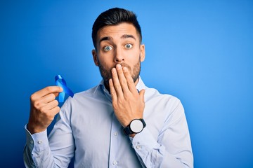 Young handsome man holding blue ribbon as prostate campaing support over blue background cover mouth with hand shocked with shame for mistake, expression of fear, scared in silence, secret concept