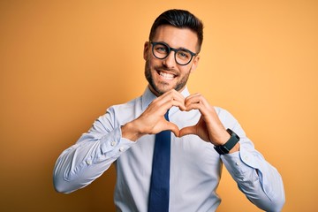 Young handsome businessman wearing tie and glasses standing over yellow background smiling in love...
