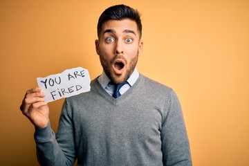 Young handsome man holding paper with you are fired message over yellow background scared in shock...