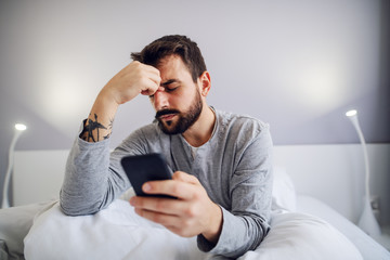 Young handsome bearded man sitting in bedroom, holding smart phone and getting nervous about...