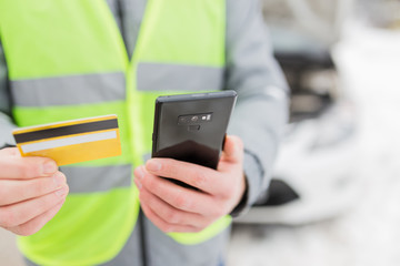 Driver holding road assistance service member card and smart phone. Broken car with opened hood in the background.