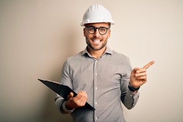Young architect inspector man wearing builder safety helmet and using clipboard very happy pointing...