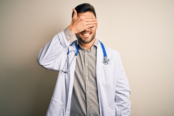 Young doctor man wearing glasses, medical white robe and stethoscope over isolated background smiling and laughing with hand on face covering eyes for surprise. Blind concept.