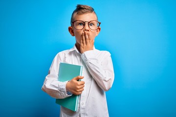 Young little caucasian student kid wearing smart glasses and holding a book from school cover mouth with hand shocked with shame for mistake, expression of fear, scared in silence, secret concept
