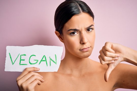 Young beautiful girl holding paper with vegan message over isolated pink background with angry face, negative sign showing dislike with thumbs down, rejection concept