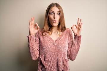 Young beautiful redhead woman wearing pink casual sweater over isolated white background looking surprised and shocked doing ok approval symbol with fingers. Crazy expression