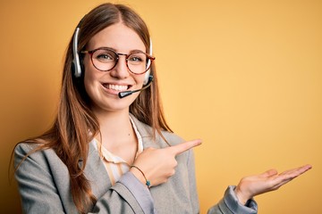 Young redhead call center agent woman overworked wearing glasses using headset amazed and smiling...