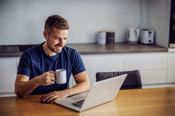 Young cheerful smiling man sitting at dinging table, holding mug with fresh morning coffee and...