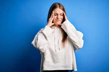 Young beautiful redhead sporty woman wearing sweatshirt over isolated blue background with hand on head for pain in head because stress. Suffering migraine.