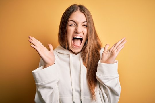 Young beautiful redhead sporty woman wearing sweatshirt over isolated yellow background celebrating crazy and amazed for success with arms raised and open eyes screaming excited. Winner concept