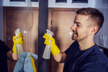 Side view of attractive man with rubber gloves on standing in front of mirror on wardrobe, spraying...