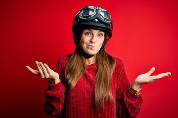 Young beautiful redhead motocyclist woman wearing moto helmet over red background clueless and confused expression with arms and hands raised. Doubt concept.