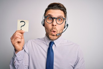 Young call center operator business man with blue eyes holding paper note with question mark scared...
