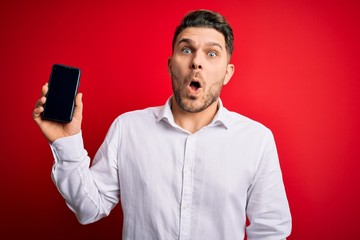 Young business man with blue eyes showing smartphone screen over red background scared in shock with a surprise face, afraid and excited with fear expression