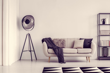 Industrial black lamp next to beige sofa with blanket and pillows and shelf with books, copy space on empty white wall