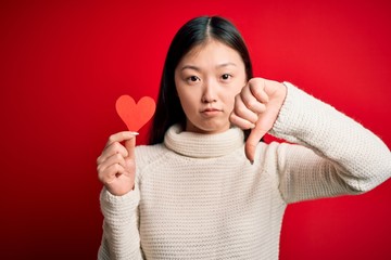 Young asian woman holding romantic red heart paper shape over red isolated background with angry...