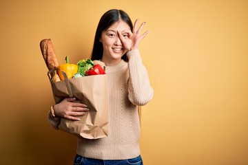 Young asian woman holding paper bag of fresh healthy groceries over yellow isolated background doing ok gesture with hand smiling, eye looking through fingers with happy face.
