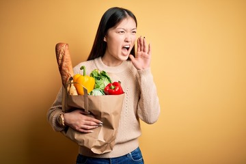 Young asian woman holding paper bag of fresh healthy groceries over yellow isolated background shouting and screaming loud to side with hand on mouth. Communication concept.