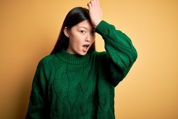 Young beautiful asian woman wearing green winter sweater over yellow isolated background surprised with hand on head for mistake, remember error. Forgot, bad memory concept.