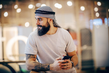 Attractive muscular fashionable arab man leaning on the railing, looking around and holding smart phone in hands. Shopping mall interior.