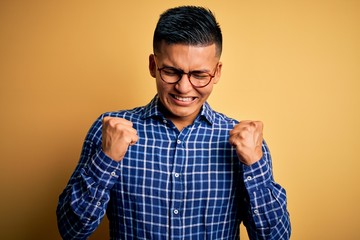 Young handsome latin man wearing casual shirt and glasses over yellow background excited for success with arms raised and eyes closed celebrating victory smiling. Winner concept.