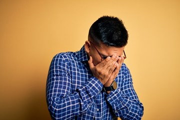 Young handsome latin man wearing casual shirt and glasses over yellow background with sad expression covering face with hands while crying. Depression concept.