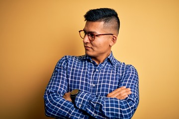 Young handsome latin man wearing casual shirt and glasses over yellow background looking to the side with arms crossed convinced and confident