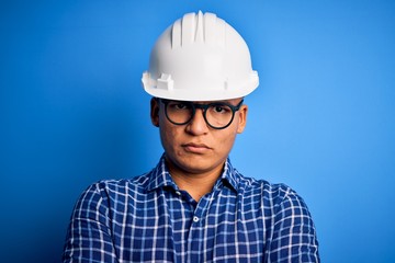 Young handsome engineer latin man wearing safety helmet over isolated blue background skeptic and...