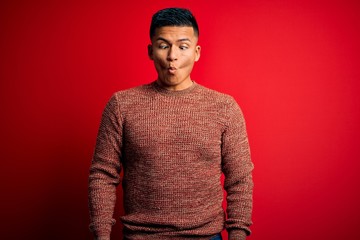 Young handsome latin man wearing casual sweater standing over red background making fish face with lips, crazy and comical gesture. Funny expression.