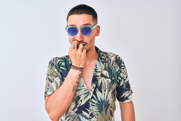 Young handsome man wearing Hawaiian sumer shirt and blue sunglasses over isolated background looking stressed and nervous with hands on mouth biting nails. Anxiety problem.